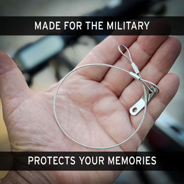 A hand holding a safety tether loop, designed for securing a GoPro Mount action camera. The background is blurred, showing parts of a bicycle. GoPro Mount action camera safety tether from Obtainium Science and Surplus.