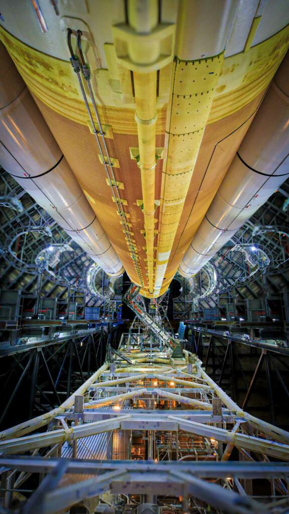 Close-up view of a large industrial cylindrical structure with yellow and beige tones, flanked by two similar cylinders, likely part of a rocket or propulsion system. The intricate support framework and piping surround the cylinders, with detailed structural components visible along the length. The setting is a high-tech facility with ambient lighting highlighting the metallic textures and engineering complexity, evoking the scale and precision of advanced aerospace or energy technology.