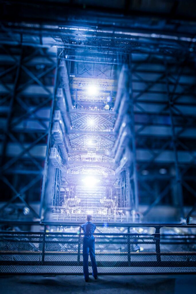 A person in a blue jumpsuit stands at a metal railing, looking up at a towering, illuminated industrial structure that extends vertically, filled with complex scaffolding and machinery. Bright lights at multiple levels cast a blue glow over the scene, creating an impression of futuristic or advanced technology, possibly related to a high-energy propulsion system or faster-than-light drive. The atmosphere is awe-inspiring, with a sense of vast scale and intense energy in the industrial setting.