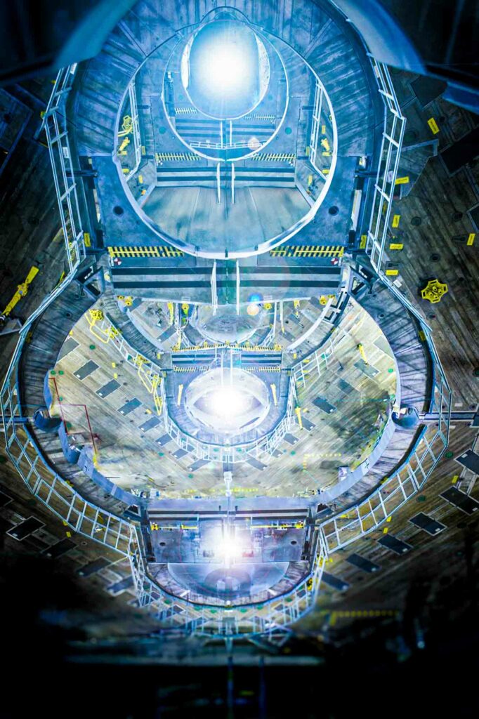 View looking down into a vast, circular industrial chamber with multiple layers and metal railings, illuminated by a series of bright, circular lights at each level. The structure features concentric platforms and safety markings in yellow, along with various metal components and reinforced walls. The chamber has a futuristic, high-tech appearance, resembling a containment or propulsion unit, evoking concepts of deep-space or energy-related technology. The arrangement creates a tunnel-like effect, drawing the eye towards the luminous center.