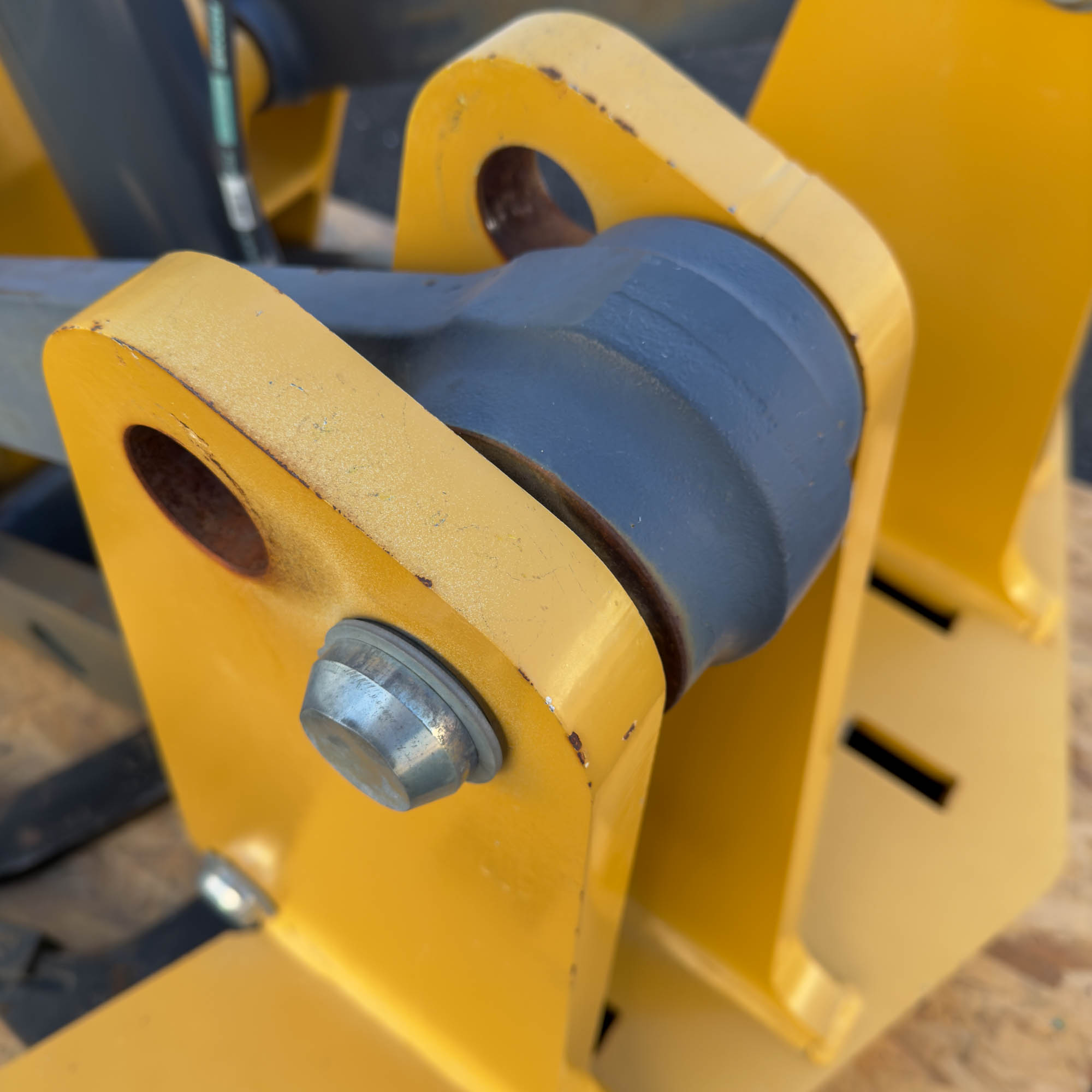 A detailed shot of the pivot point on the yellow Front Mount Scarifier for John Deere Motor Graders from Obtainium Science and Surplus, focusing on a steel roller.