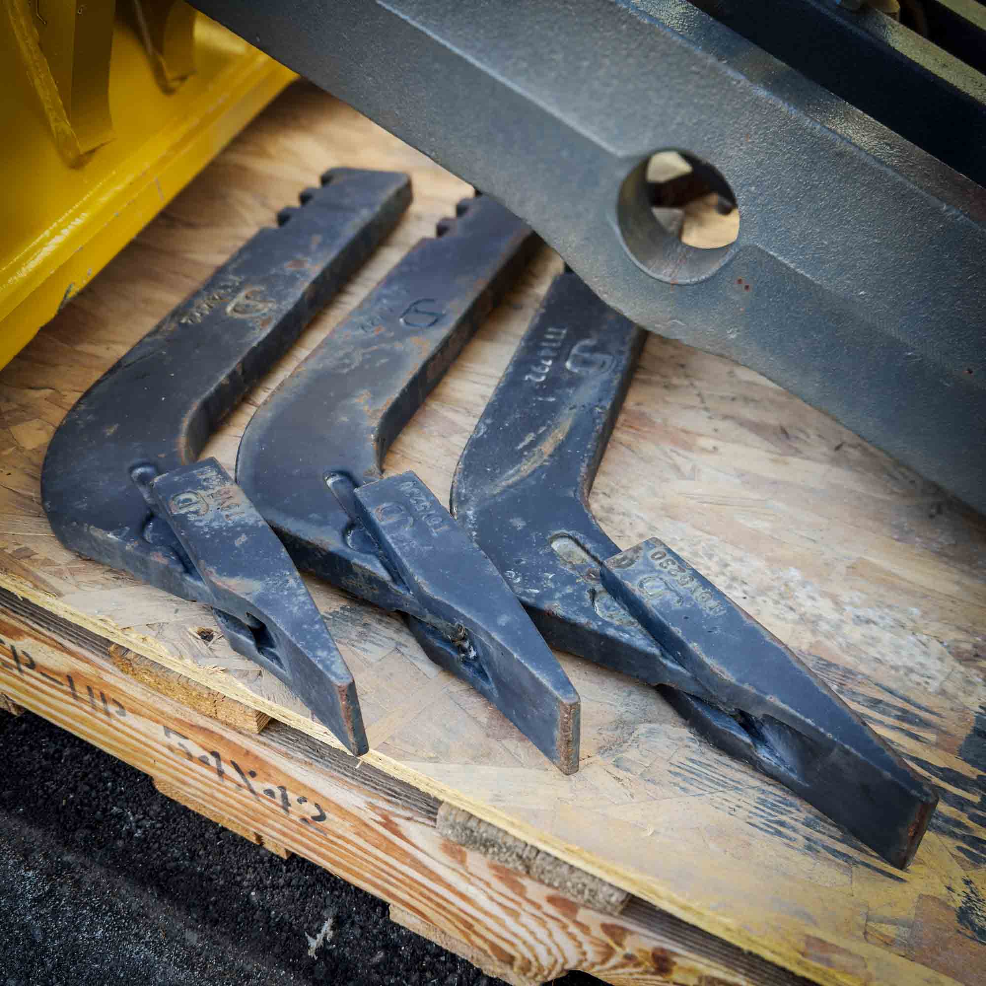 Three scarifier tooth and shank assemblies for the Front Mount Scarifier for John Deere Motor Graders from Obtainium Science and Surplus, placed on a wooden pallet.