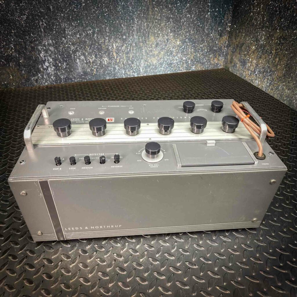 A top-angle view of the Leeds & Northrup Model 7556 Guarded Potentiometer from Obtainium Science and Surplus. The device has a sturdy metal body, black knobs, and a row of input jacks, with a power cord resting on the side.