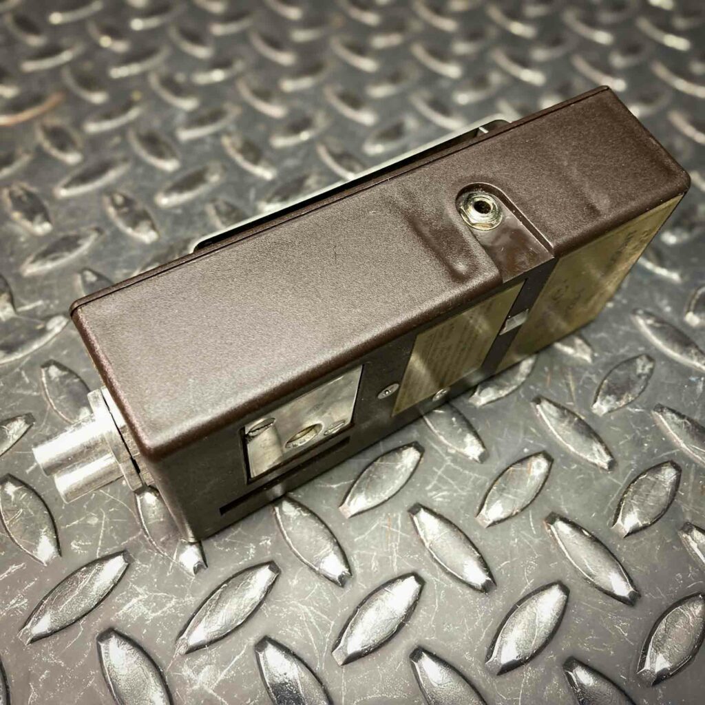 A side view of the SKC low-flow personal air sampler model 222 from Obtainium Science and Surplus, showing the dark brown casing and a small opening near the edge. The device is resting on a textured metal surface.