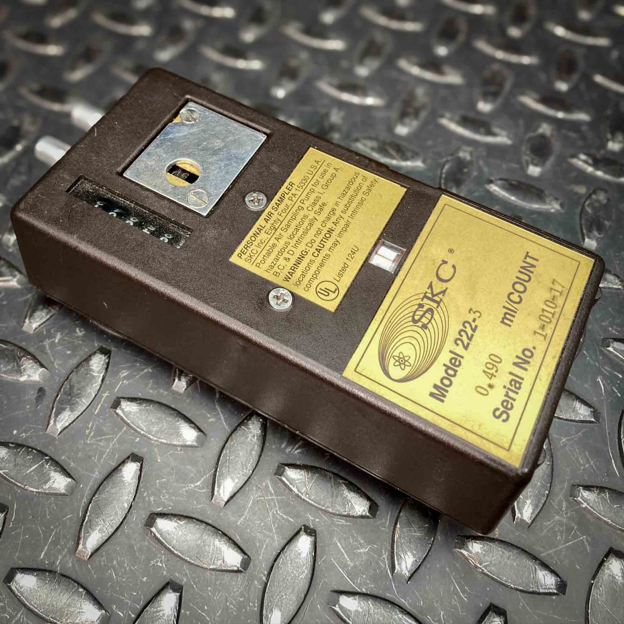 Another angled view of the SKC low-flow personal air sampler model 222 from Obtainium Science and Surplus, displaying the yellow label, counter window, and adjustment dial. The dark brown casing contrasts with the textured metal surface beneath it.