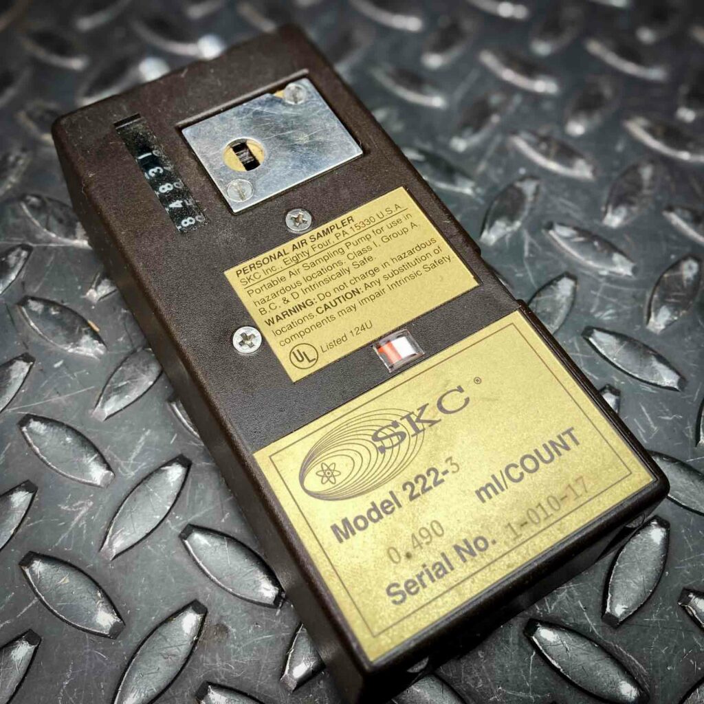 An SKC low-flow personal air sampler model 222 from Obtainium Science and Surplus, placed on a textured metal surface. The device is rectangular, dark brown, with a yellow label displaying model details and a serial number. A small window and control features are visible on the top.