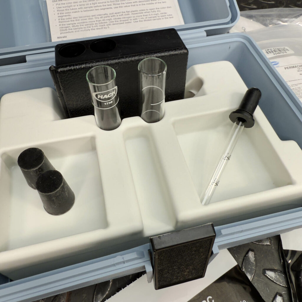 Close-up of the open Hach Water Test Kit for Nitrites and Nitrates from Obtainium Science and Surplus. Inside the blue case, there are two glass test tubes, black caps, and a plastic dropper placed in a white foam insert. The background shows part of the instruction manual.