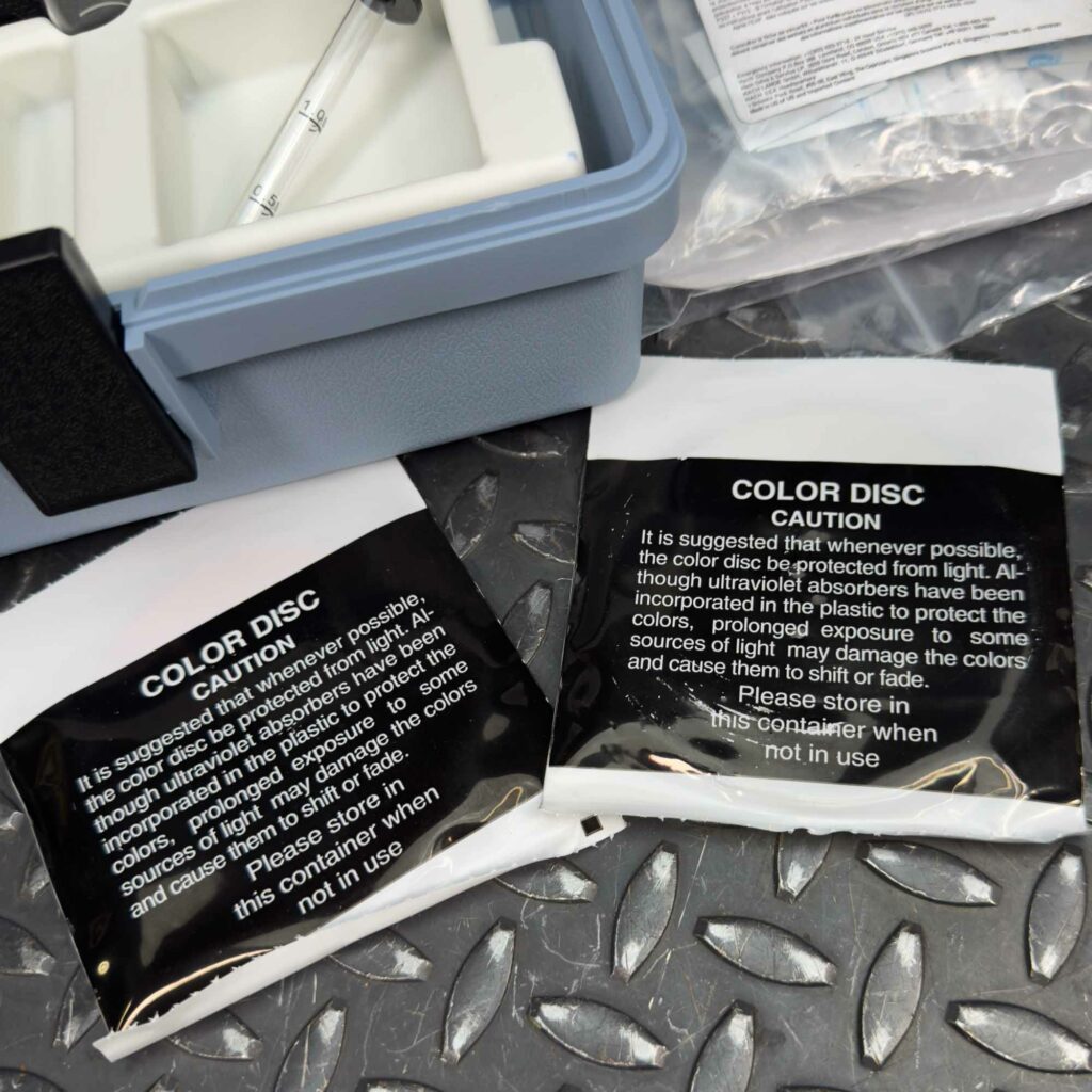 Two small reagent packets labeled "COLOR DBC" next to the open Hach Water Test Kit for Nitrites and Nitrates from Obtainium Science and Surplus. The packets contain chemical reagents for testing, with instructions and safety warnings printed on the packaging.