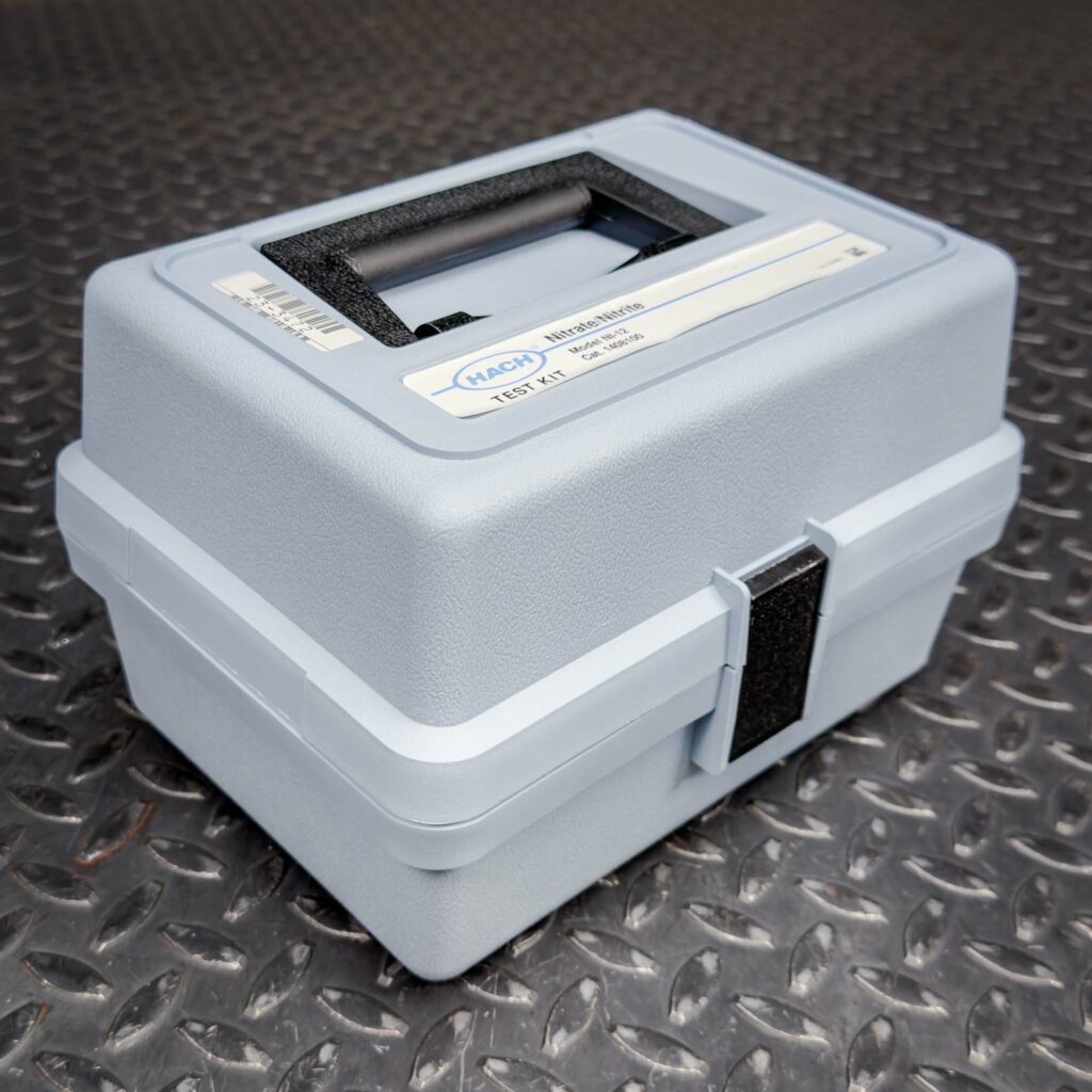 A closed blue plastic carrying case for the Hach Water Test Kit for Nitrites and Nitrates from Obtainium Science and Surplus, resting on a textured metal surface. The case has a black handle and a secure latch.