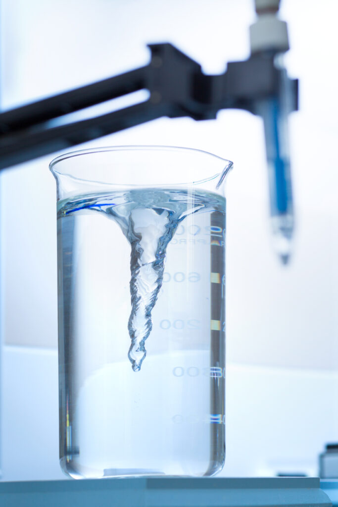 A laboratory beaker filled with clear liquid sits on a magnetic stirrer, creating a visible vortex in the center due to the rapid spinning of a magnetic stir bar. The beaker has measurement markings and is illuminated by bright lighting, highlighting the swirling motion of the liquid. In the blurred background, a laboratory clamp holds a pH probe or similar scientific instrument.